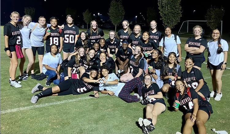 girls on a flag football team standing up and sitting down on the field smiling for a photo. most are in black jerseys. 